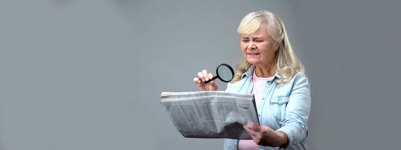 senior with magnifiying glass reading paper 1280×480
