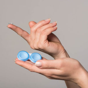 Soft contact lens and blue container in female hands on gray bac