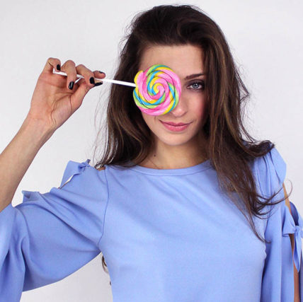 brunette woman wearing blue dress holding large lollypop over right eye
