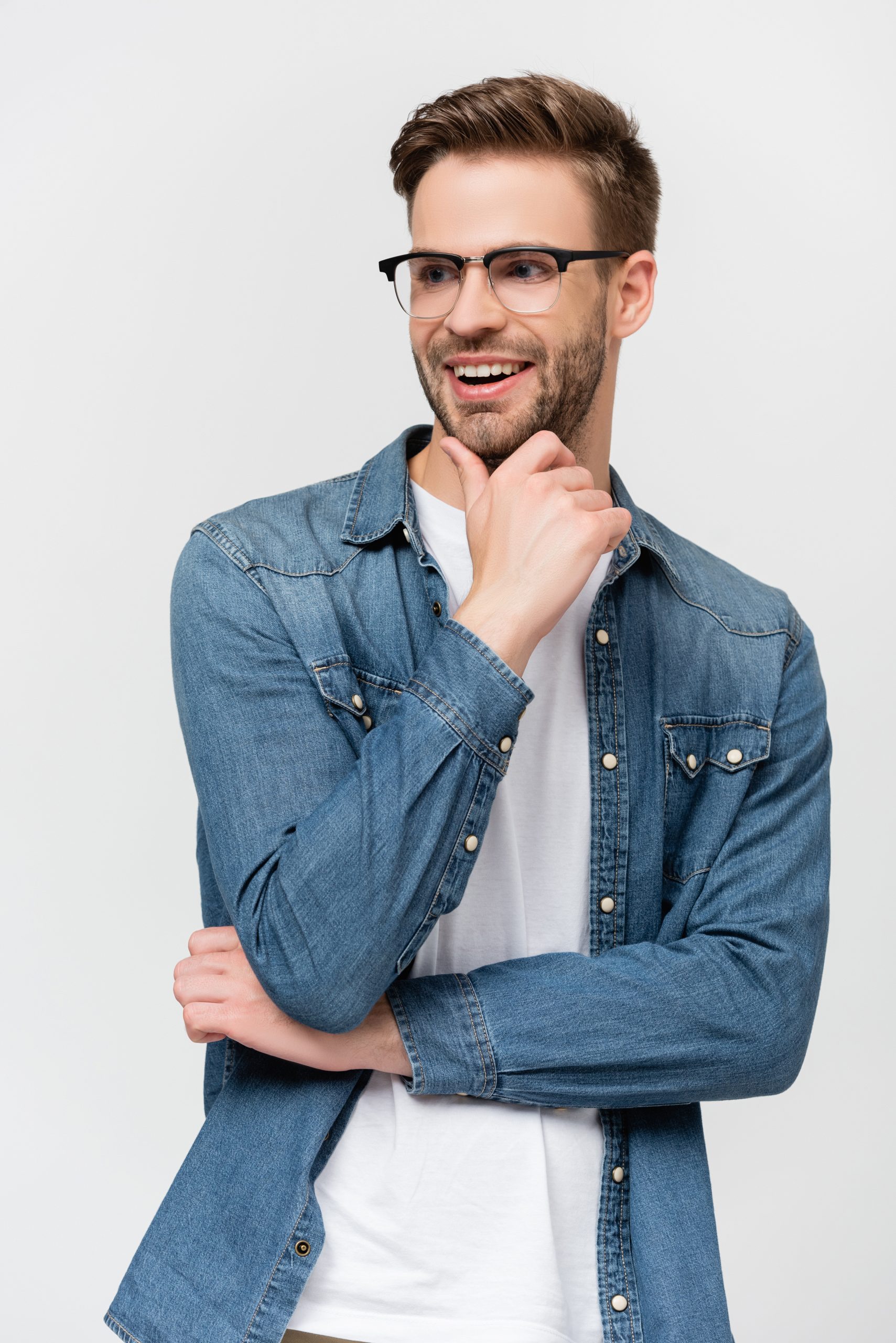 Young man in eyeglasses smiling while touching chin isolated on grey