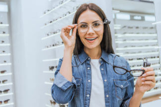 cheerful young woman choosing eyeglasses and looking at camera in ophthalmic shop