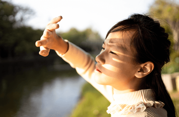 Child holding a hand up in front of their face while looking directly into the sun and frowning
