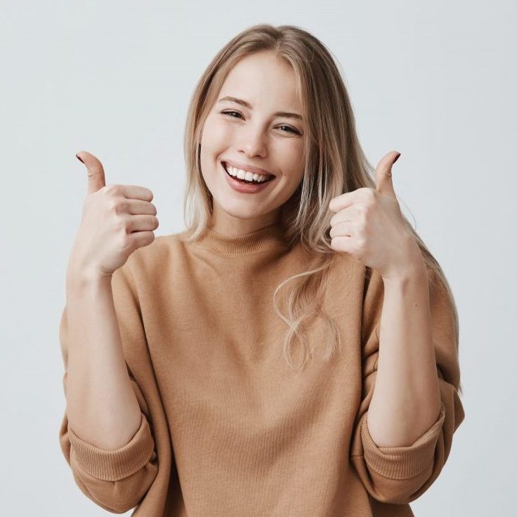 Portrait of fair haired beautiful female student or customer with broad smile, looking at the camera with happy expression, showing thumbs up with both hands, achieving study goals. Body language