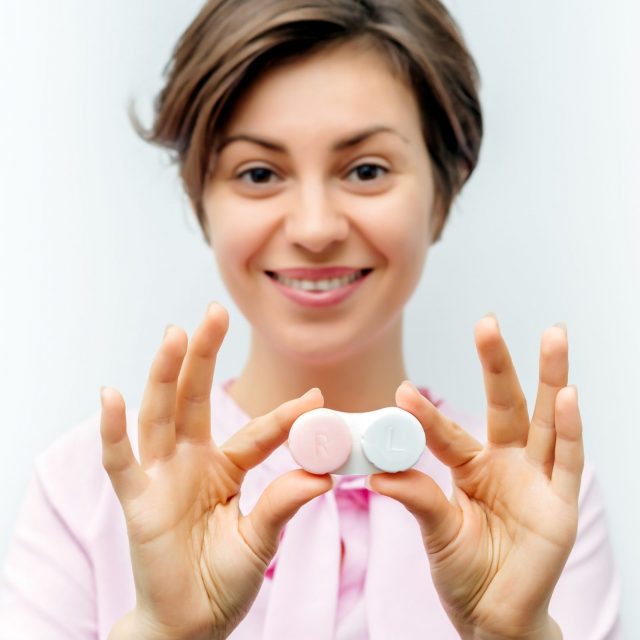 Smiling woman in pink blouse holding a box of contact lenses, shows them to the camera. focus on the hands.