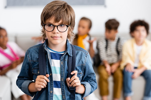 schoolboy in eyeglasses holding backpack in classroom