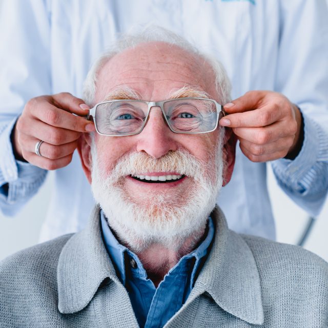 Doctor fitting glasses on cheerful aged male patient: cropped photo