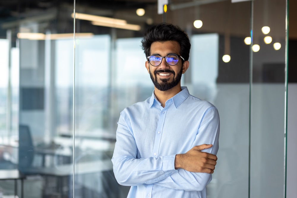 Portrait of a young Indian male designer, engineer, architect who is wearing glasses and a blue shirt smiling standing in the office and looking at the camera