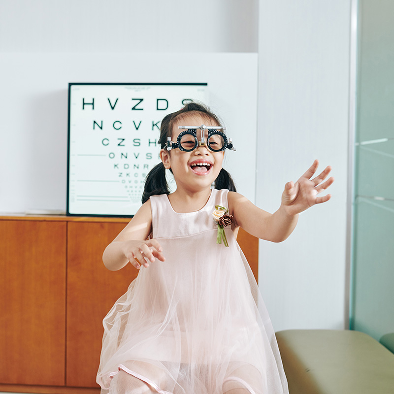 Cheerful laughing little Vietnamese girl in test glasses in office of pediatric optometrist