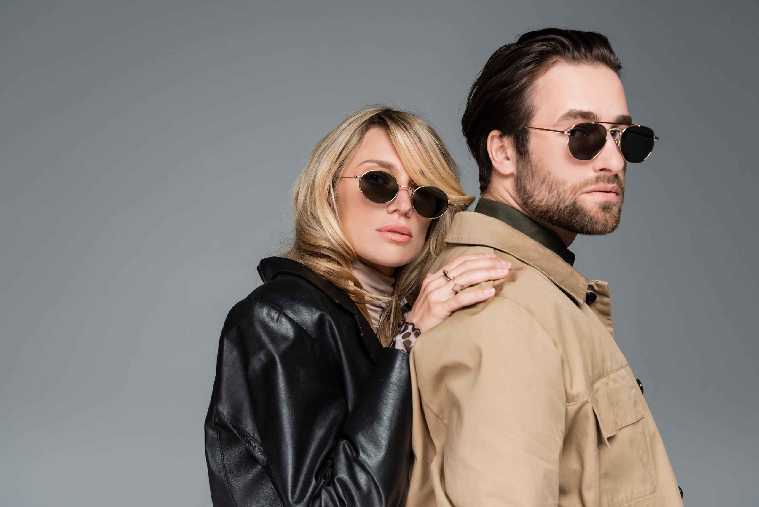 stylish blonde woman in sunglasses and leather jacket leaning on shoulders on bearded man isolated on grey