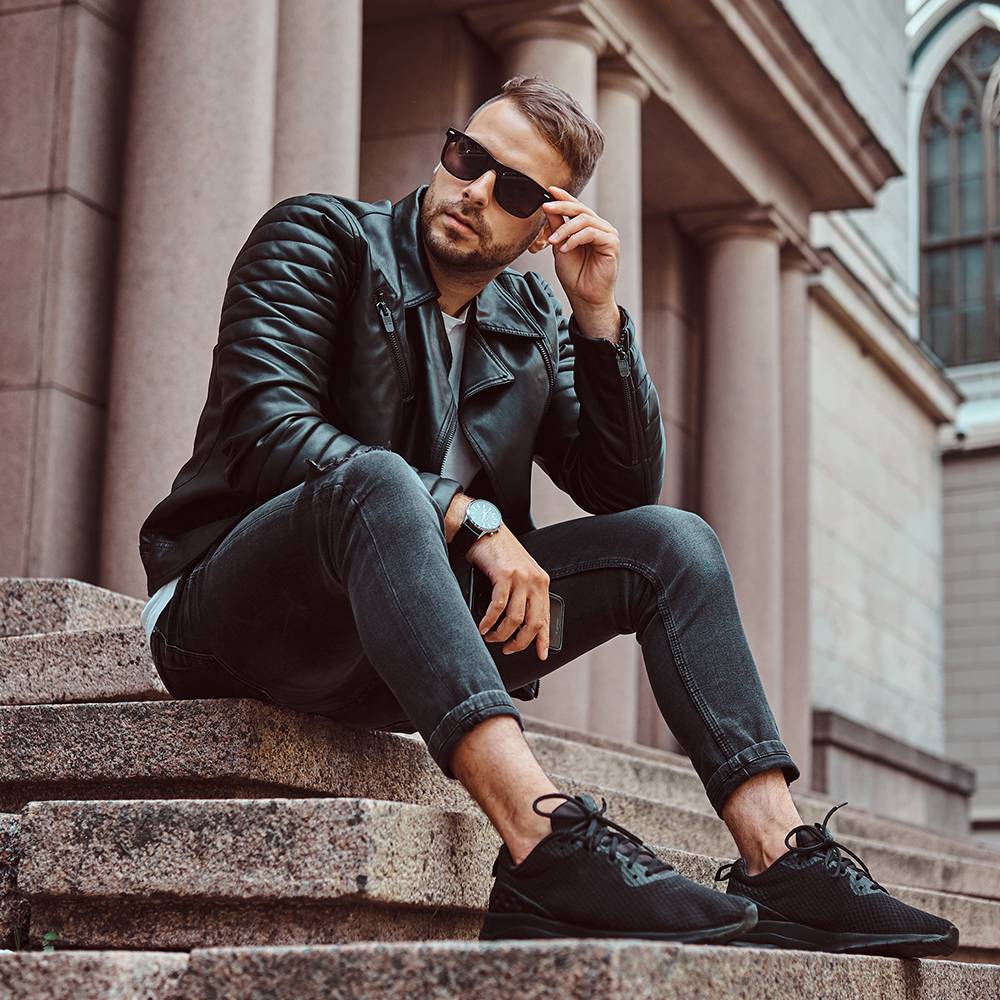 Fashionable guy dressed in a black jacket and jeans holds the smartphone sitting on steps against an old building in Europe.