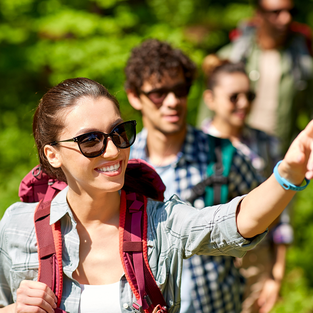 travel, tourism, hike and people concept—woman showing something to group of friends walking with backpacks in forest