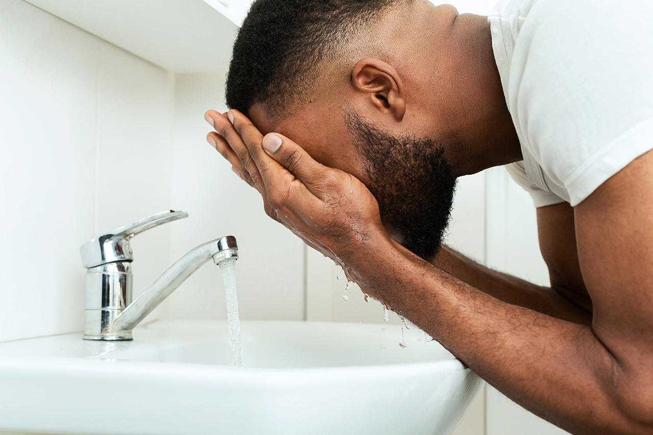 Black man washing his face, splashing water at sink in bathroom