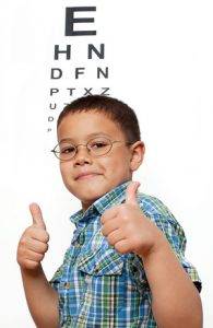 Cute little boywith glasses smiling and giving thumbs up sign, in front of eye chart in the background. Children at the eye doctor optometrist theme.