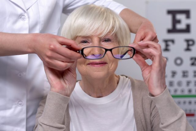 Optician wearing elderly female patient new glasses, vision trea