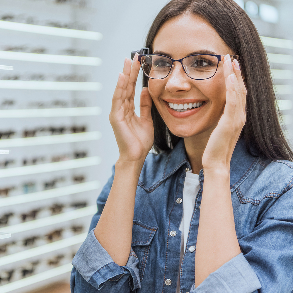 partial view of smiling woman choosing eyeglasses and looking at mirror in optica
